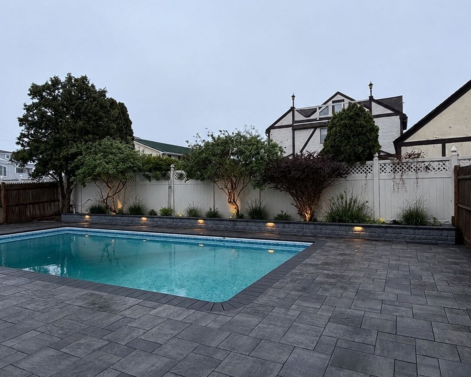 Paver pool deck lit at dusk along a white privacy fence