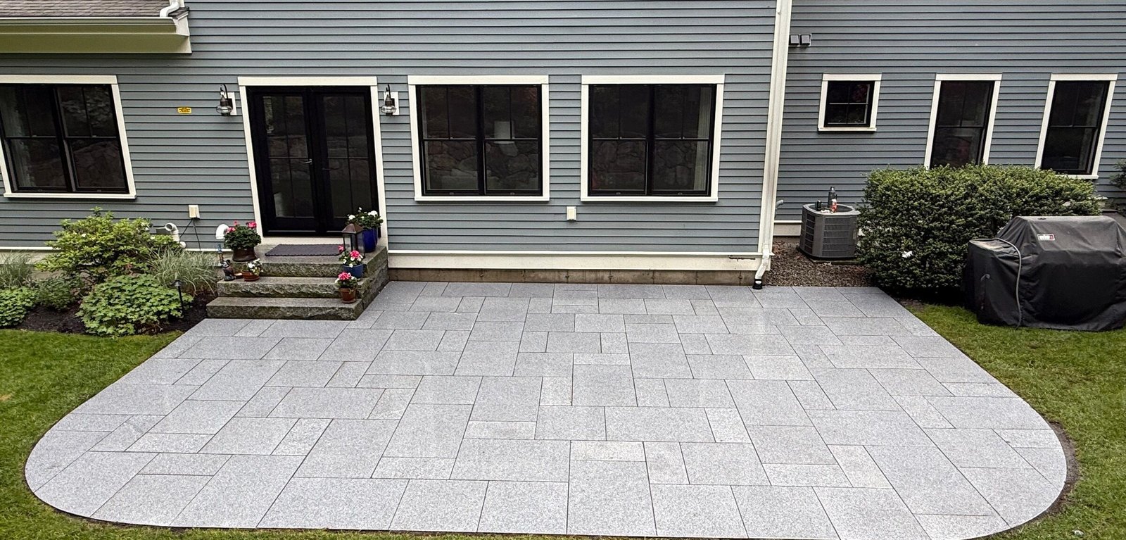 Light granite paver patio curving into a manicured lawn behind a gray home